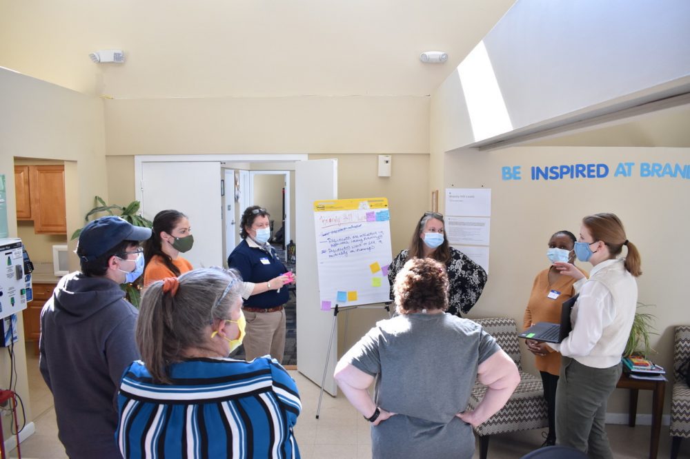 A workshop scene: Eight people stand in a circle, all masked (except possibly the one whose back is to the camera). A pony-tailed woman on the right holds up a laptop so the others can see it, and there's a chalk-talk pad at the far end with writing on it. Trauma-informed community building