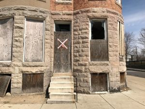 A vacant rowhouse in Baltimore sits boarded up, with a large white 'X' spray painted on the piece of plywood covering the door.