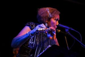 A screengrab of Margaux Simmons, co-organizer of Music City, playing the flute, standing at a microphone with theatrical lighting highlighting her face and arms.