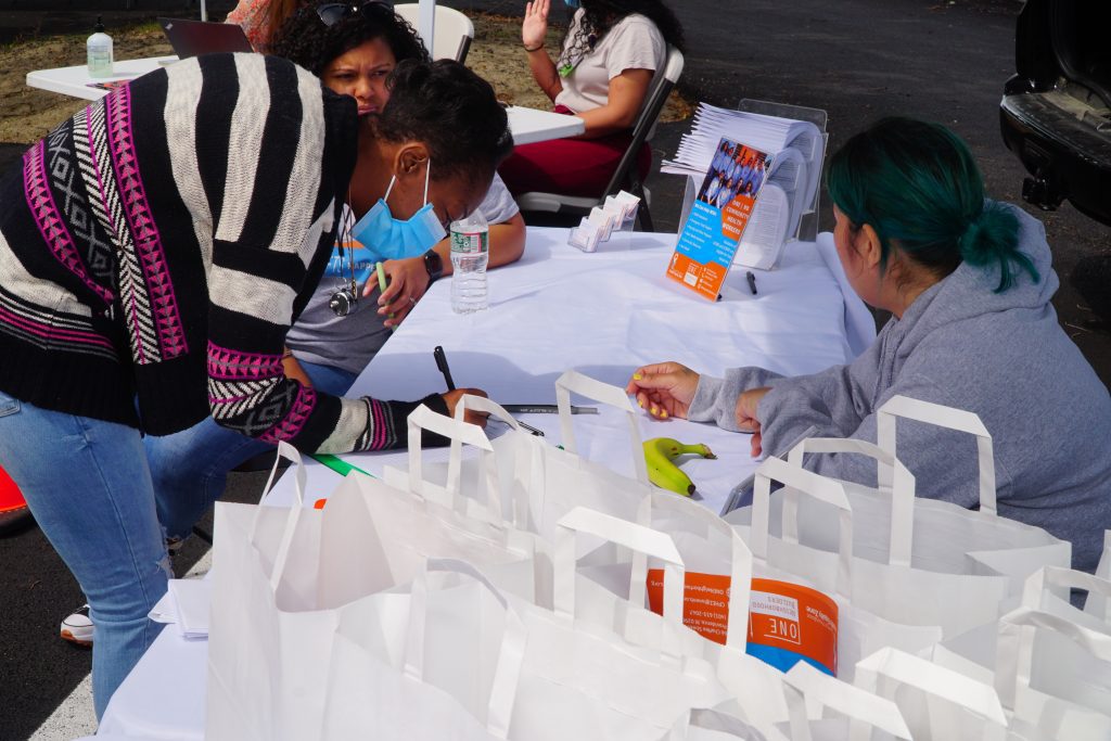 A woman in a striped sweater and wearing a surgical mask leans over a table where she is writing something. Another woman is seated on the other side of the table, a staffer from a community development organization. Near her are white paper bags that contain brochures.