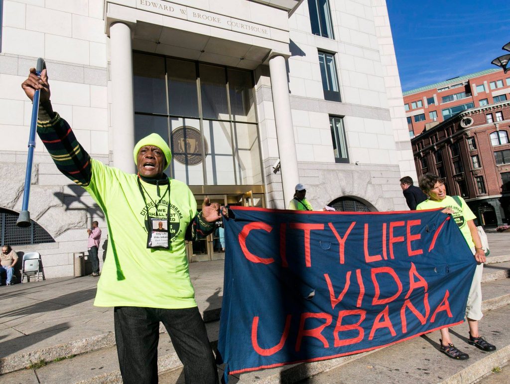 In front of the Edward W. Brooke Courthouse, two people stand holding a banner reading "City Life/Vida Urbana." CLVU volunteer assist vulnerable tenants facing eviction in court unassisted.