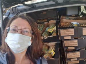 A selfie of a woman, wearing a surgical mask, in front of an open car or truck cargo area showing pineapples stacked high.
