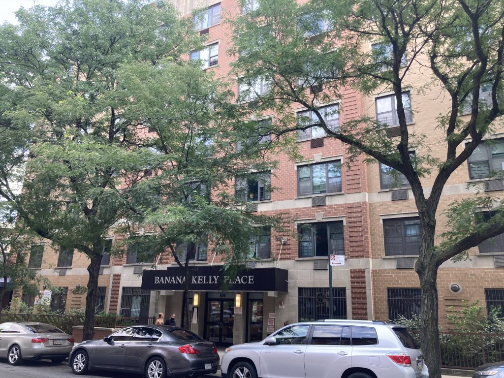 A view from the street of Banana Kelly Place in the Bronx. The multistory brick building has a dark awning with "Banana Kelly Place" in capital latters. Three cars are parked in front of the building and leafy trees line the sidewalk.