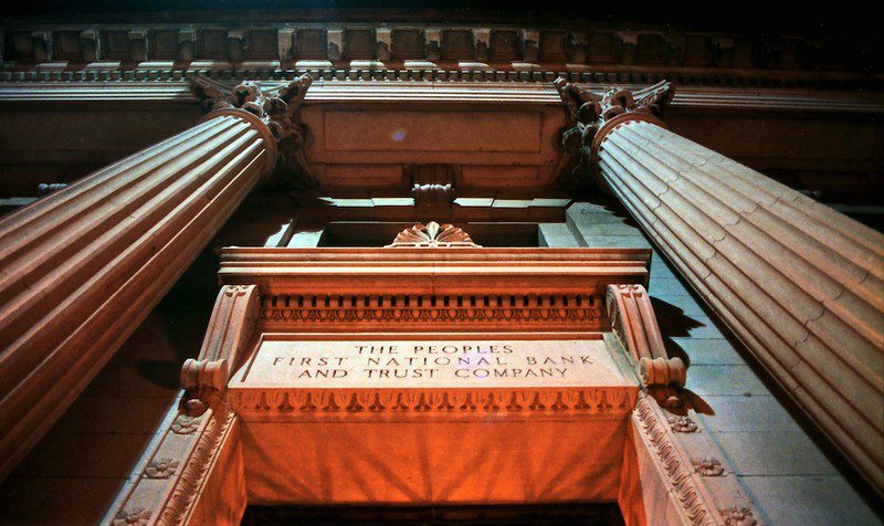 A ground-level view looking up at the entrance to The Peoples First National Bank and Trust Company. The name is carved over the door and topped by a ledge and flanked by fluted columns of stone. 