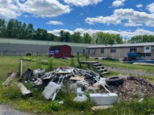 A large pile of debris from a demolished mobile home contrasts with the blue sky, fluffy clouds, and green trees in the distance. In the middle distance is an intact mobile home.