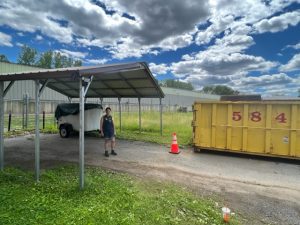 Under a blue sky with fluffy clouds, a large yellow dumpster sits by the carport of a mobile home. Someone is standing under the carport roof.