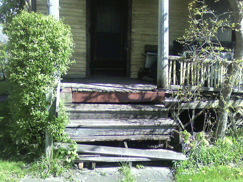 An exterior view of the unpainted wooden front steps of a dilapidated house. The lowest step is hanging off the riser and the middle steps look very worn. There has been an attempt at repair on the top riser; it's a red-painted board.