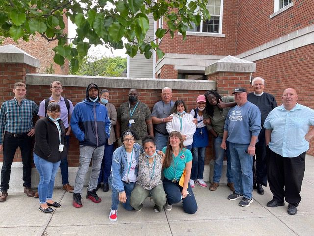 Exterior photo of Joseph's House workers and supporters. Sixteen people stand in front of a brick wall. They are all ages; some are wearing green lanyards, others not.