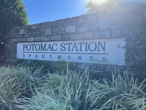 A stone wall bears a sign saying "Potomac Station Apartments." In front of the wall are some tufty ornamental grasses.