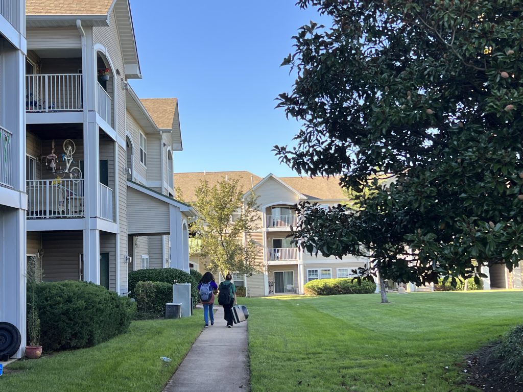Exterior of The Fields at Cascades, an apartment complex, with two people on a walkway in front of a three story building with balconies. Ahead of them is a similar-looking building and at right is a tree in full foliage. Tenants here are facing mid-lease rent increases.