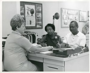 A blac-and-white photo showing a white woman behind a desk, while an African American couple fill out application forms to become residents at Co-op City.