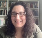 Close-up of author Annemarie Sammartino. She has curly brown hair, wears glasses, and is seen in front of a tall bookcase filled with books.