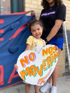 A little girl in a yellow T-shirt holds a hand-painted heart-shaped sign that reads "No Nos Moverán." Behind her is a woman, partly visible, holding one end of a blue banner.