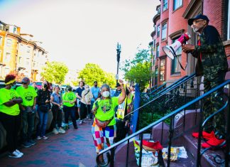 A sidewalk view of a front stoop where a man stands holding a bullhorn. Lined up on the sidewalk in front of him is a large group of people, many wearing CLVU's bright yellow-green T-shirts.