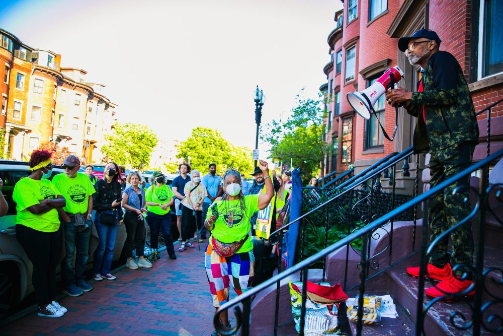 A sidewalk view of a front stoop where a man stands holding a bullhorn. Lined up on the sidewalk in front of him is a large group of people, many wearing CLVU's bright yellow-green T-shirts.