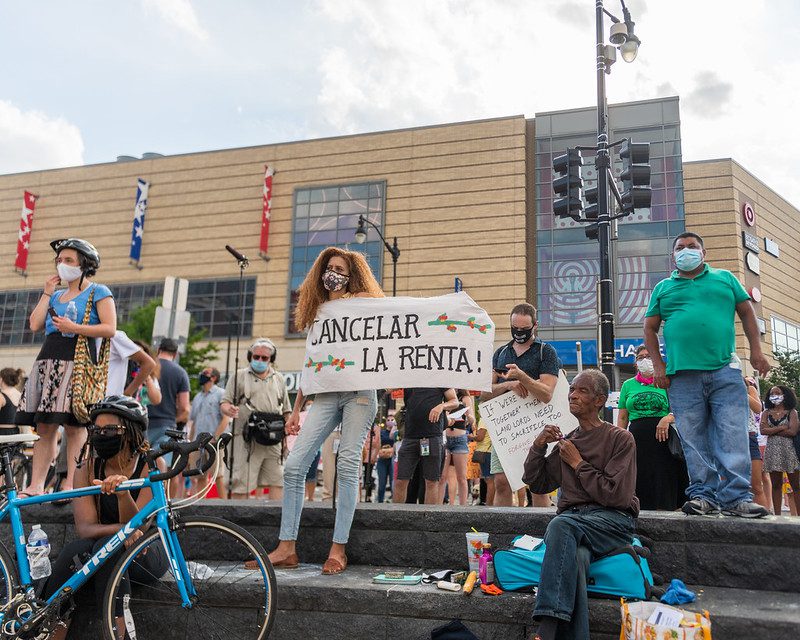 A group of people, many masked, stand outside on some steps. One woman in the middle with curly auburn hair holds a sign that reads "Cancelar la Renta," translated to "Cancel the Rent."