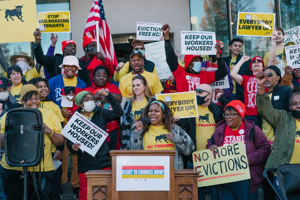 A large group of people, most holding signs, at a rally in support of right to counsel for tenants. Some of the signs say "Keep our workers housed!" "Stop Railroading Tenants," and "No More Evictions." Many of the people are wearing yellow T-shirts under their jackets; the shirts say "KC Tenants."