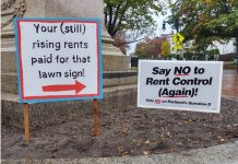 Two campaign signs stand together near the base of a monument in Portland, Maine. The sign at right reads "Say NO to Rent Control (Again)!" The sign on the left has a red arrow pointing to the other sign, and the text says: "Your (still) rising rents paid for that lawn sign!"