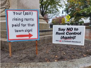 Two campaign signs stand together near the base of a monument in Portland, Maine. The sign at right reads "Say NO to Rent Control (Again)!" The sign on the left has a red arrow pointing to the other sign, and the text says: "Your (still) rising rents paid for that lawn sign!"