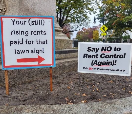 Two campaign signs stand together near the base of a monument in Portland, Maine. The sign at right reads "Say NO to Rent Control (Again)!" The sign on the left has a red arrow pointing to the other sign, and the text says: "Your (still) rising rents paid for that lawn sign!"