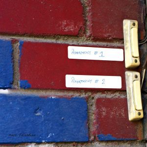 A close-up view of two electronic doorbells on a brick wall. The bricks are painted red and blue in a pattern that the viewer is too close to to see. The doorbells, which are grubby-looking, have paper stickers next to them reading Apartment #1 or Apartment #2.