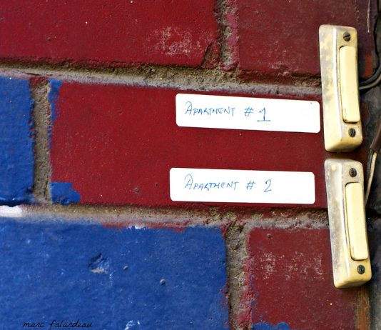 A close-up view of two electronic doorbells on a brick wall. The bricks are painted red and blue in a pattern that the viewer is too close to to see. The doorbells, which are grubby-looking, have paper stickers next to them reading Apartment #1 or Apartment #2.