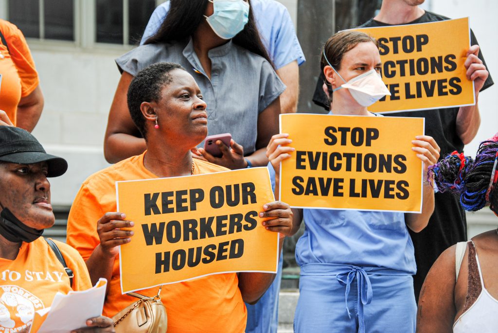 A group of people stand on steps during a press conference, but the faces of the ones in back are cropped out. Three in the group are holding up orange signs that say "Keep our workers housed" or "Stop evictions save lives." In the center of the photo are a Black woman in an orange T-shirt and a white woman in blue.