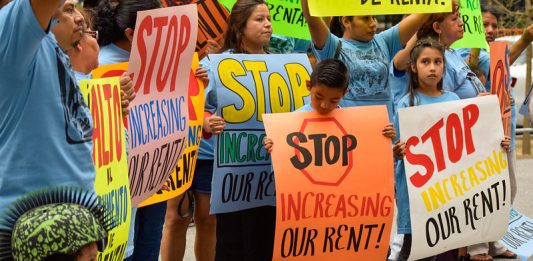 A group of adults and children stand with colorful orange, yellow and blue signs. One sign reads, "Stop increasing our Rent."