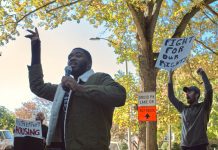 One man holds a microphone and raises his other hand while speaking outside, and behind him, a person holds a white and black sign.