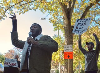 One man holds a microphone and raises his other hand while speaking outside, and behind him, a person holds a white and black sign.