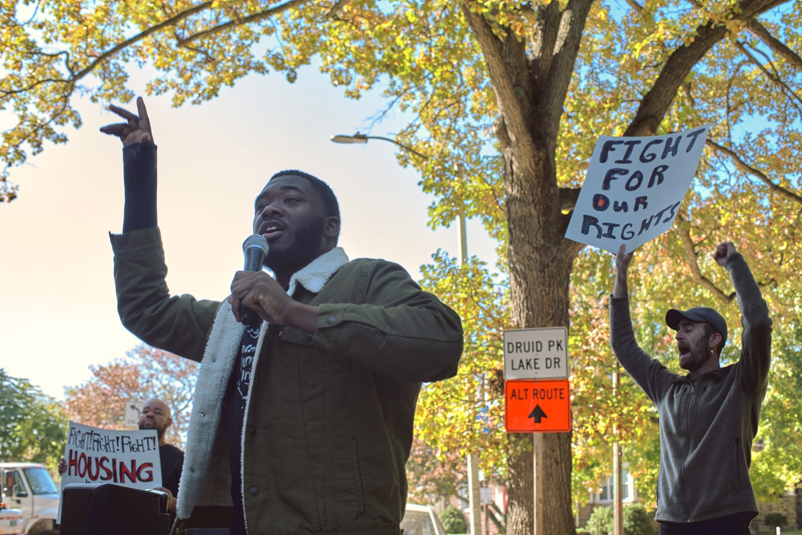 One man holds a microphone and raises his other hand while speaking outside, and behind him, a person holds a white and black sign.