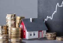 Stock photo shows a small toy house partly surrounded by stacks of coins. Near it on a blackboard is drawn a jagged upward line, symbolizing rising real estate prices