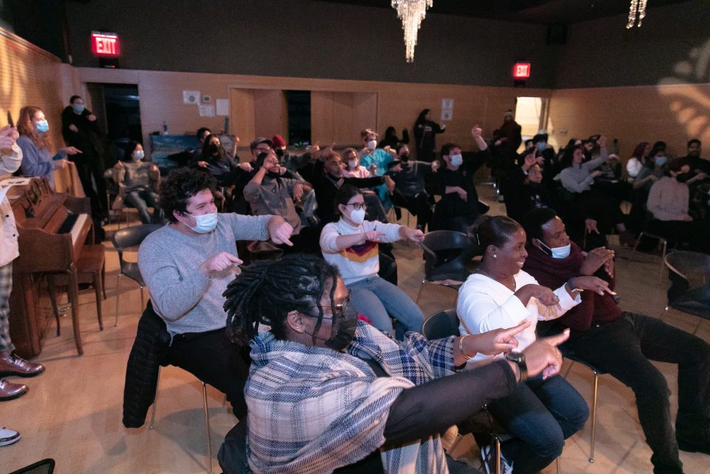 Several dozen people, most seated but some leaning on the wall, and most wearing masks, hold their arms in the air, fingers pointing. Some are smiling.