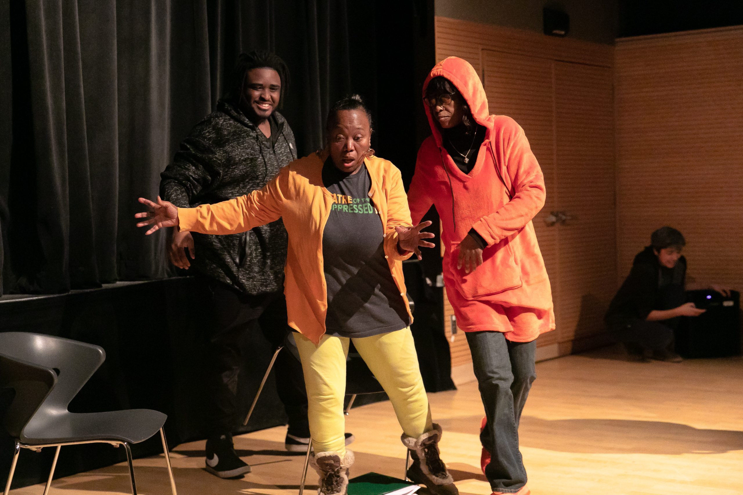On a spotlit theater stage, three people enact a scene. One is in back, in dark colors. The other two are dressed in bright peaches and yellows, and one is holding her arm out.