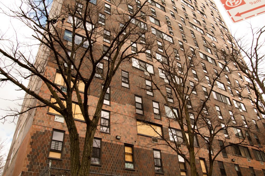 A view from the street looking up at a 19-story apartment building. Bare wintertime trees make it possible to see boarded-up windows on the lower floors and thick soot from a fire surrounding one window.