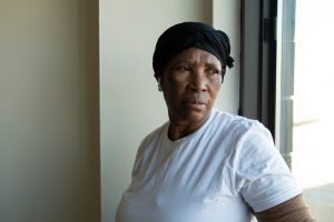 An African American woman of 66 with a somber expression stands by the window of her fire-damaged apartment, looking over her shoulder toward the light. No furnishings are visible in the photo.