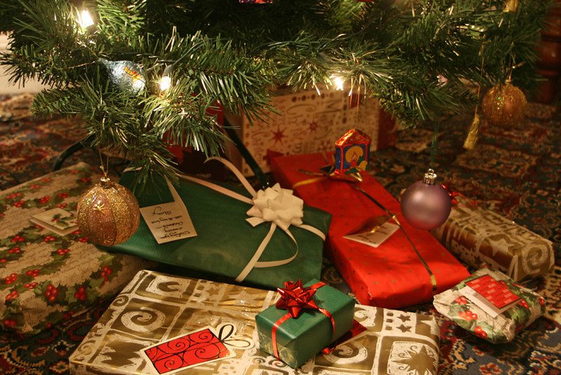 A pile of presents wrapped in red and green paper with bows, beneath a Christmas tree.