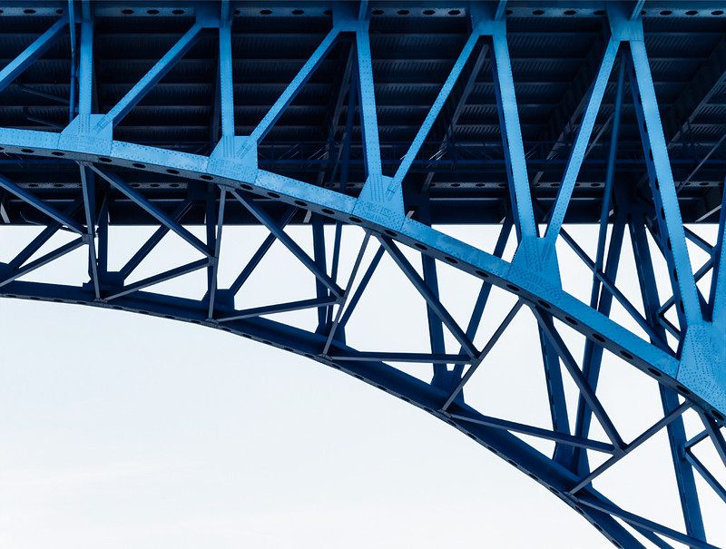 A view from below of the girders supporting a bridge. They're a bright blue, in contrast to the black underside of the roadway and the white sky.