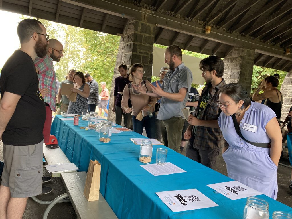 A group of folks standing surround a long table that has a teal tablecloth.