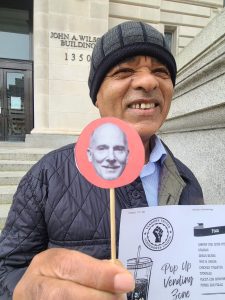 A man in a knit cap and gray quilted jacket stands on the steps of a municipal office building. He could be either smiling or squinting in the light. He's holding a dowel on which is attached a small headshot photo of a man (looks like a large lollipop). He is also holding a paper that's partly visible. It has the Vendors United?Vendedores Unidos logo and says "Pop Up Vending Zone."