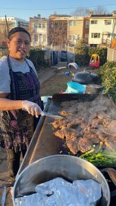 An outdoor sunny-day photo of a woman wearing a print apron over her clothes. She's holding tongs and cooking at a large griddle nearly covered with slices of meat and a mound of scallions. In the background of the small yard she's in are children's riding toys and a barbecue. Beyond that are the backs of houses on the next street.