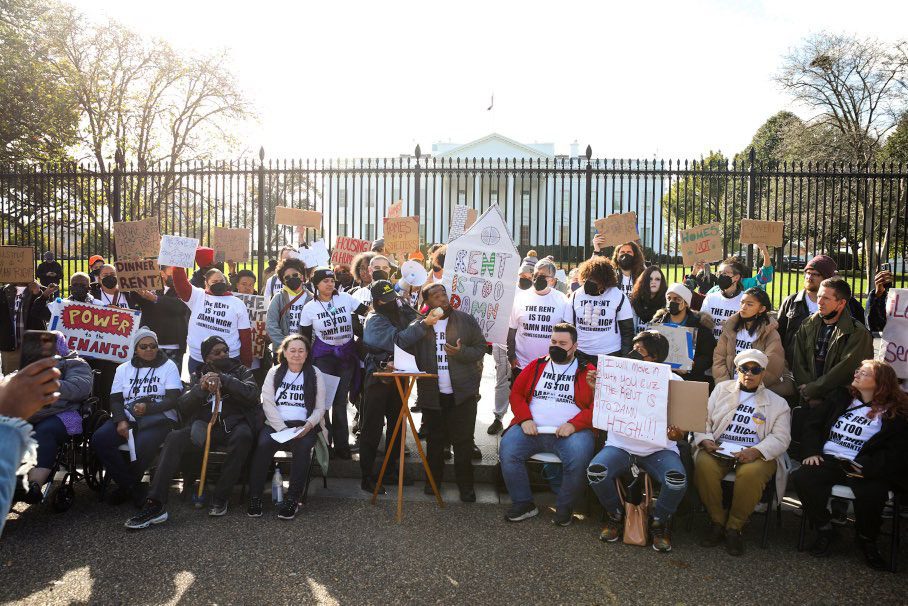 A group of 30 or 40 people standing in front of the fence near the White House. They're wearing T-shirts that say "The rent is too damn high" and holding signs on whiteboard and cardboard.