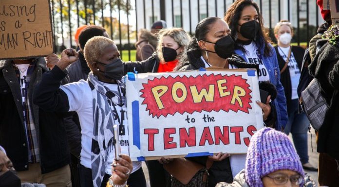 Two women in a crowd of protesters hold a big hand-lettered sign that says "Power to the tenants." Other people near and behind them are also holding signs.