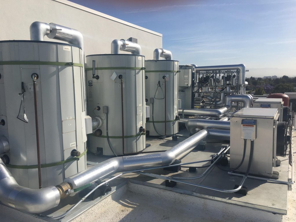 A row of large gray cylindrical water storage tanks on a roof. Behind them are heat pumps and other technical apparatus.