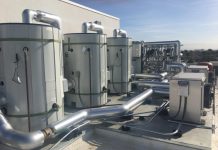 A row of large gray cylindrical water storage tanks on a roof. Behind them are heat pumps and other technical apparatus.