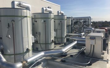 A row of large gray cylindrical water storage tanks on a roof. Behind them are heat pumps and other technical apparatus.