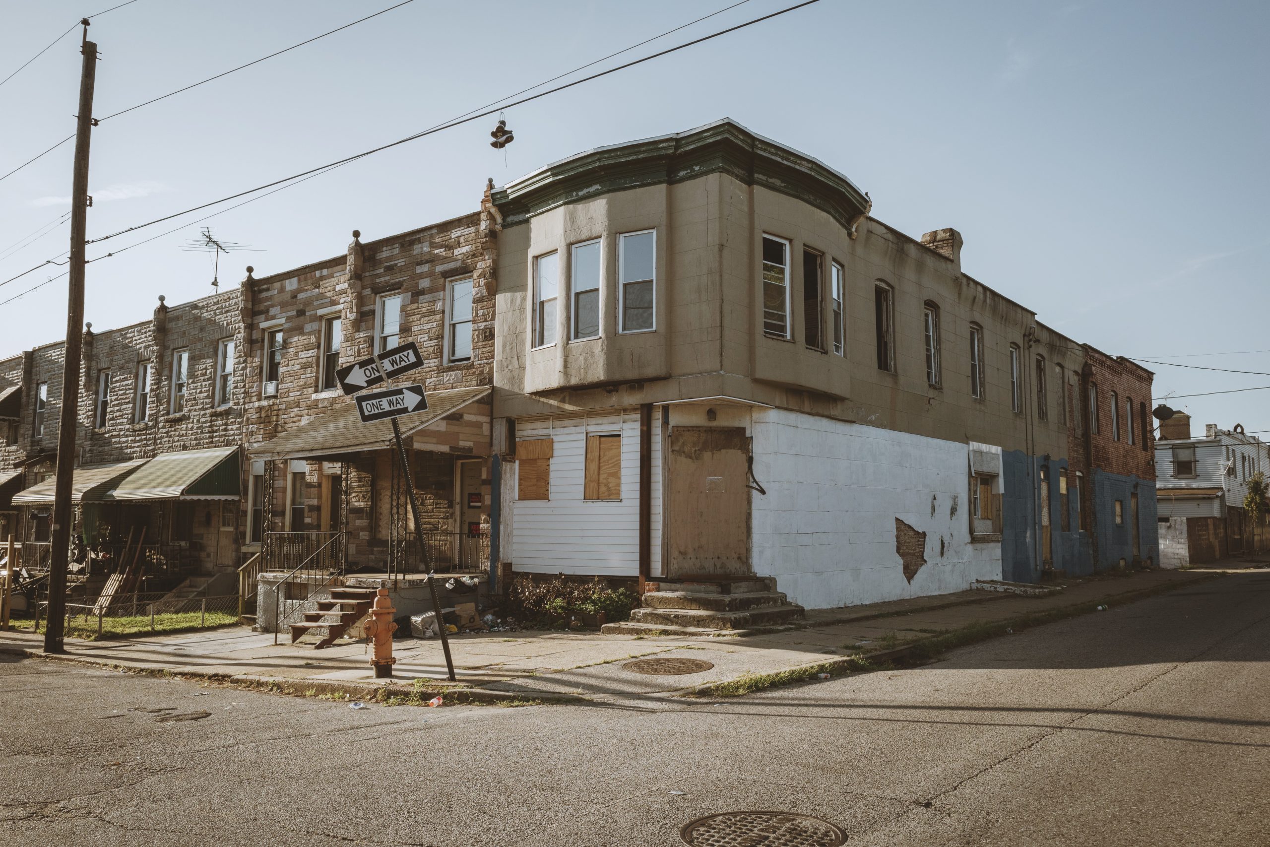 View from across the intersection of a rundown-looking corner in Baltimore, all two-story rowhouses. Some windows are boarded up. There are no cars or people in the scene.