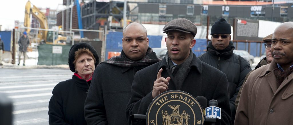 Six people in winter coats and hats stand across the street from a construction site. At the center, a young man in a cloth cap stands at a podium gesturing as he speaks. 