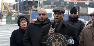 Six people in winter coats and hats stand across the street from a construction site. At the center, a young man in a cloth cap stands at a podium gesturing as he speaks.