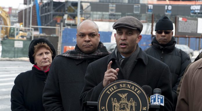 Six people in winter coats and hats stand across the street from a construction site. At the center, a young man in a cloth cap stands at a podium gesturing as he speaks.